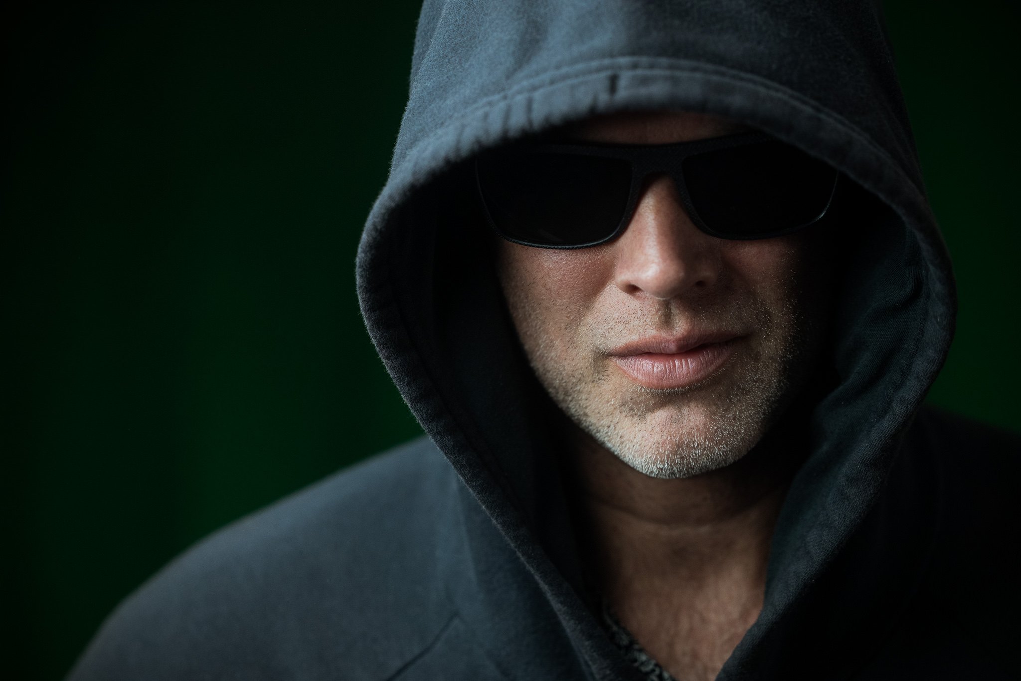 Retrato de una persona con capucha y lentes oscuros sobre un fondo verde.