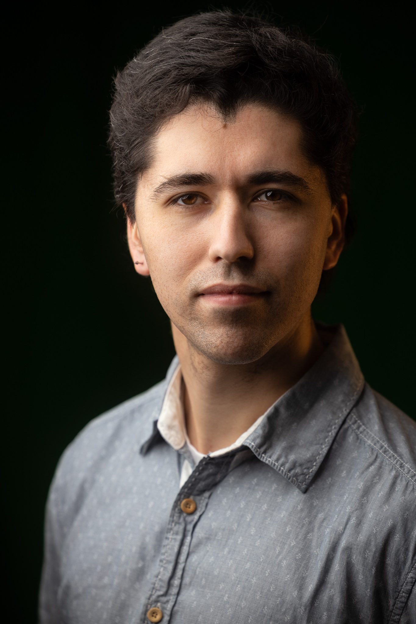 Retrato frontal de una persona con luz suave sobre un fondo verde oscuro.